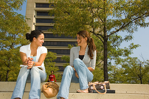 Two students sitting on steps talking and laughing with campous buildings in background.