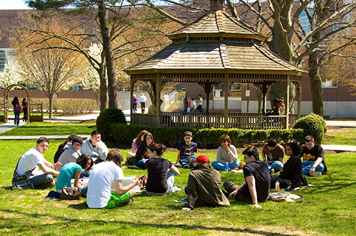Students talking and sitting in circle on grass in front of gazebo on campus with buildings in the background out